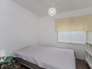 A small, single-bed room with a desk and chair, a window with blinds, and a hanging light fixture.