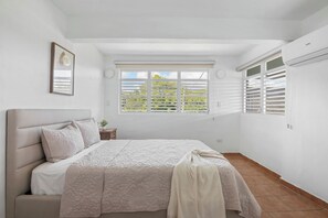 Ein Schlafzimmer mit Bett, einem Fenster mit Jalousien, einer Wand-Klimaanlage und einem kleinen Nachttisch.