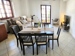 Ein Wohnzimmer mit einer Couch, einem Esstisch mit Stühlen, einem Fernseher und einem Fenster mit Vorhängen.
