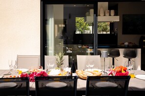 An outdoor dining area with a table set for two, featuring a variety of fruits, a bottle, and glasses. - La Badoumba · Heated Pool & Jacuzzi (Saleilles)
