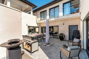 A modern outdoor patio with a grill, seating, and a view of the building's interior. - La Badoumba · Heated Pool & Jacuzzi (Saleilles)