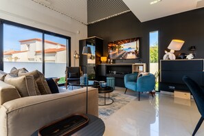 A modern living room with a sofa, a coffee table, and a TV. There is a balcony with a view of buildings. - La Badoumba · Heated Pool & Jacuzzi (Saleilles)