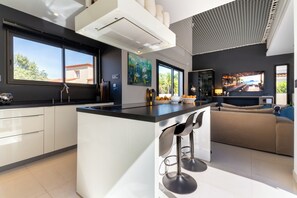 A modern kitchen with a large island, bar stools, and a TV. - La Badoumba · Heated Pool & Jacuzzi (Saleilles)