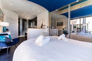 A modern hotel room with a large bed, a desk, a chair, and a bathroom with a glass shower. - La Badoumba · Heated Pool & Jacuzzi (Saleilles)