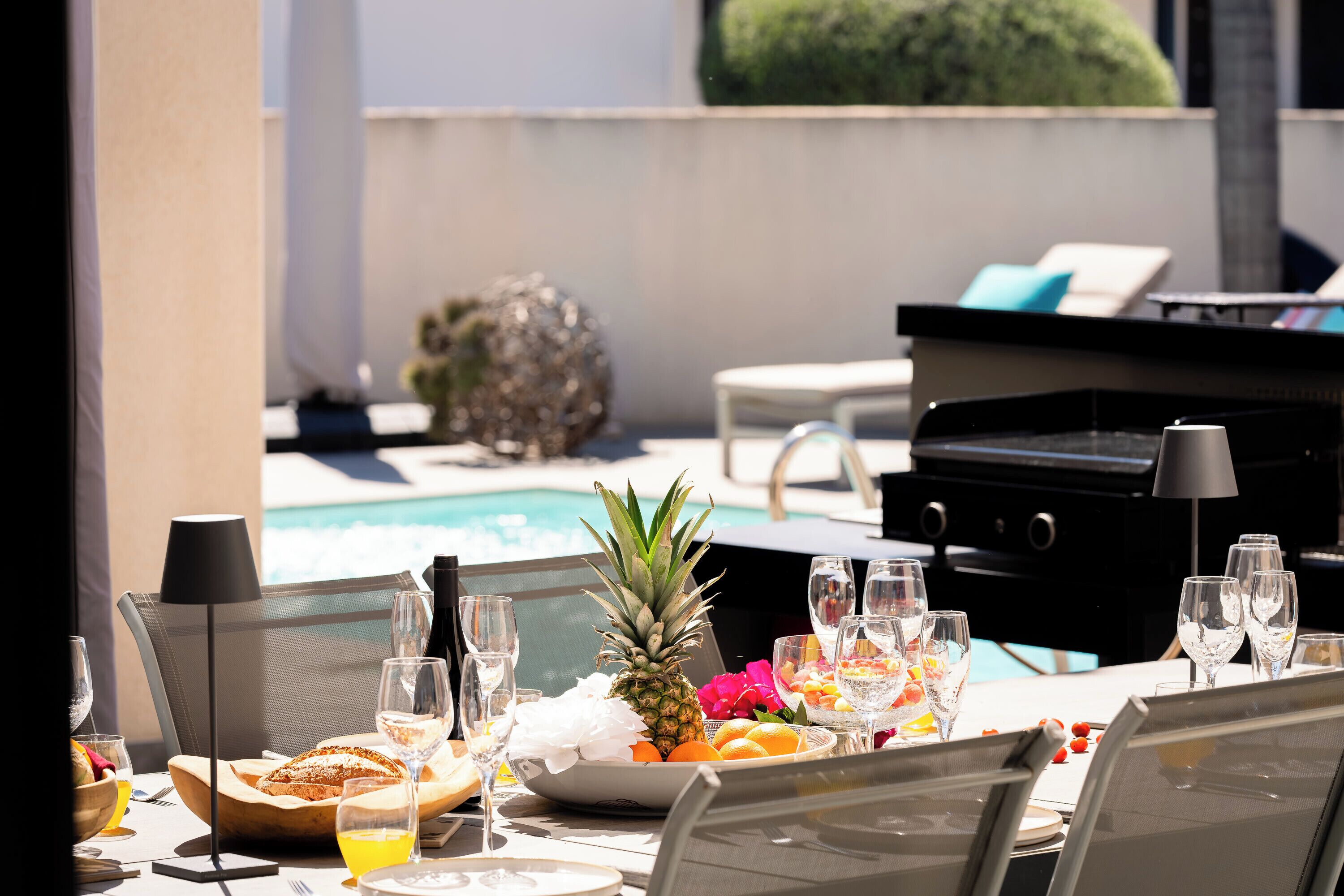 An outdoor dining area with a pool view, a grill, and a table set with fruits and drinks.