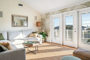 A living room with a sofa, coffee table, and a large mirror. There is a door leading to an outdoor area with a view. - Tropical 6BR Sandbridge w/ Pool + Beach Access (Virginia Beach)