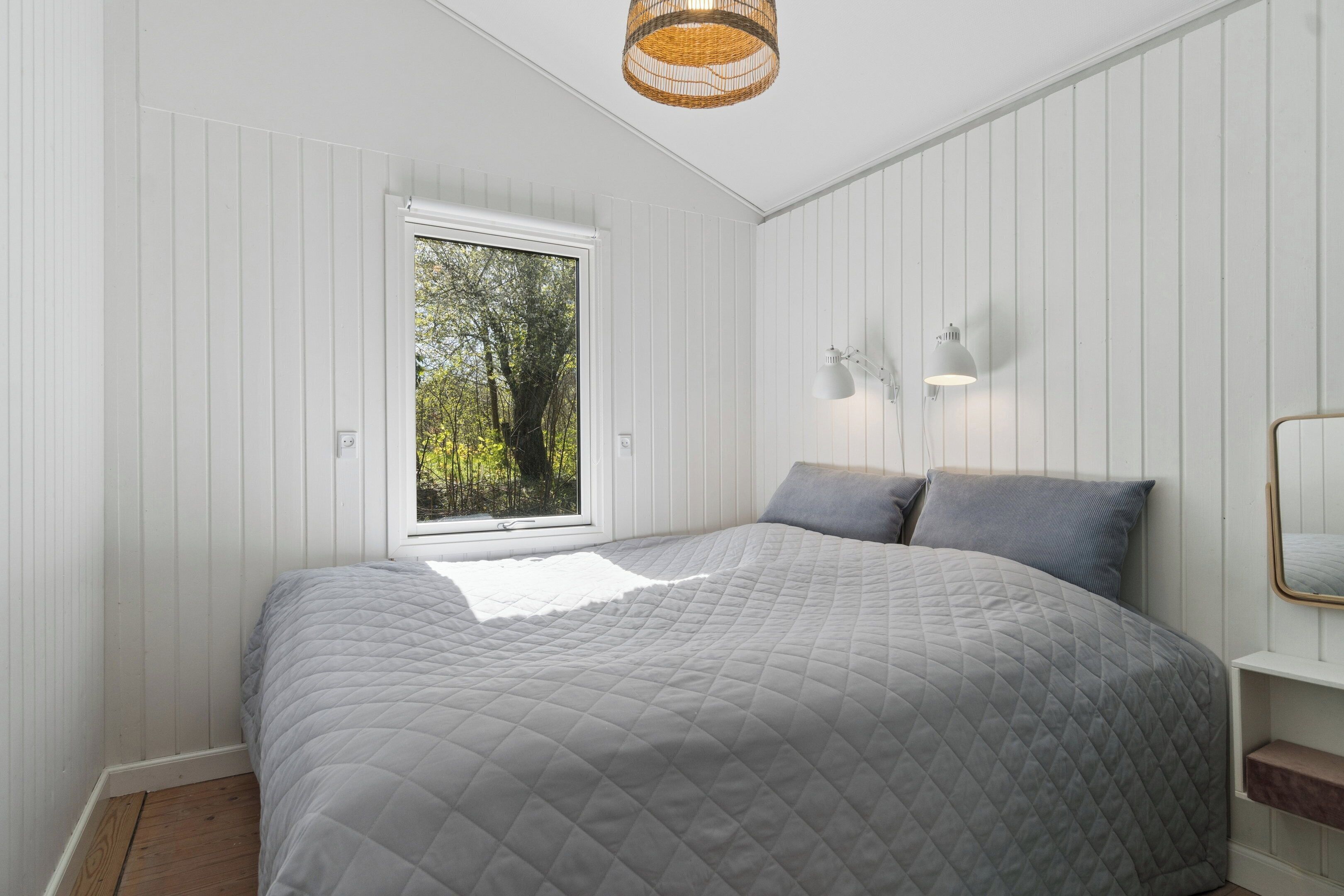 A bedroom with a large bed, a window with a view of trees, a wall-mounted lamp, and a ceiling light.