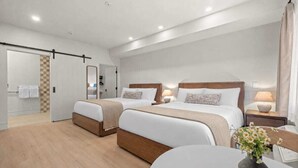 Une chambre d’hôtel avec deux lits, une salle de bain avec un sol à carreaux, un miroir et un vase de fleurs posé sur une table.