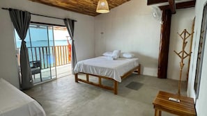 Un dormitorio con cama, mesita de noche de madera, balcón con vista al mar y un amplio ventanal con cortinas.