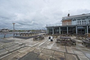 Eine Uferterrasse mit Picknicktischen, einem Mülleimer und einem Gebäude mit großen Fenstern.