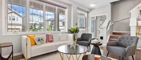 Uma sala de estar ampla com um sofá branco, duas poltronas, uma mesa de centro e uma janela grande com vista para as casas vizinhas.