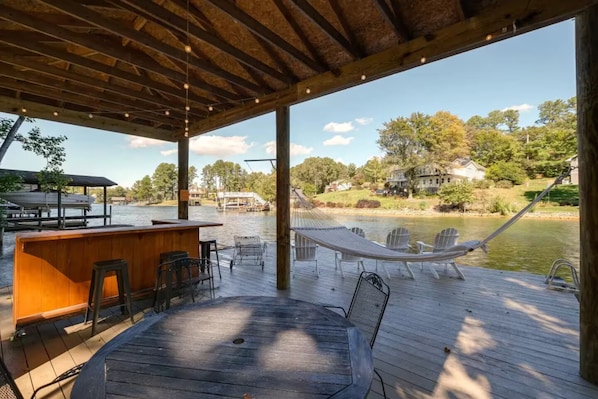 A wooden deck with a table and chairs, a hammock, and a bar area overlooking a lake. - Lakefront Retreat w/ Private Dock, Firepit + Kayaks Sunset Views (Moneta)