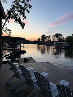 A wooden deck with Adirondack chairs, a dock extending into calm water, and a dog near the dock. - Lakefront Retreat w/ Private Dock, Firepit + Kayaks Sunset Views (Moneta)