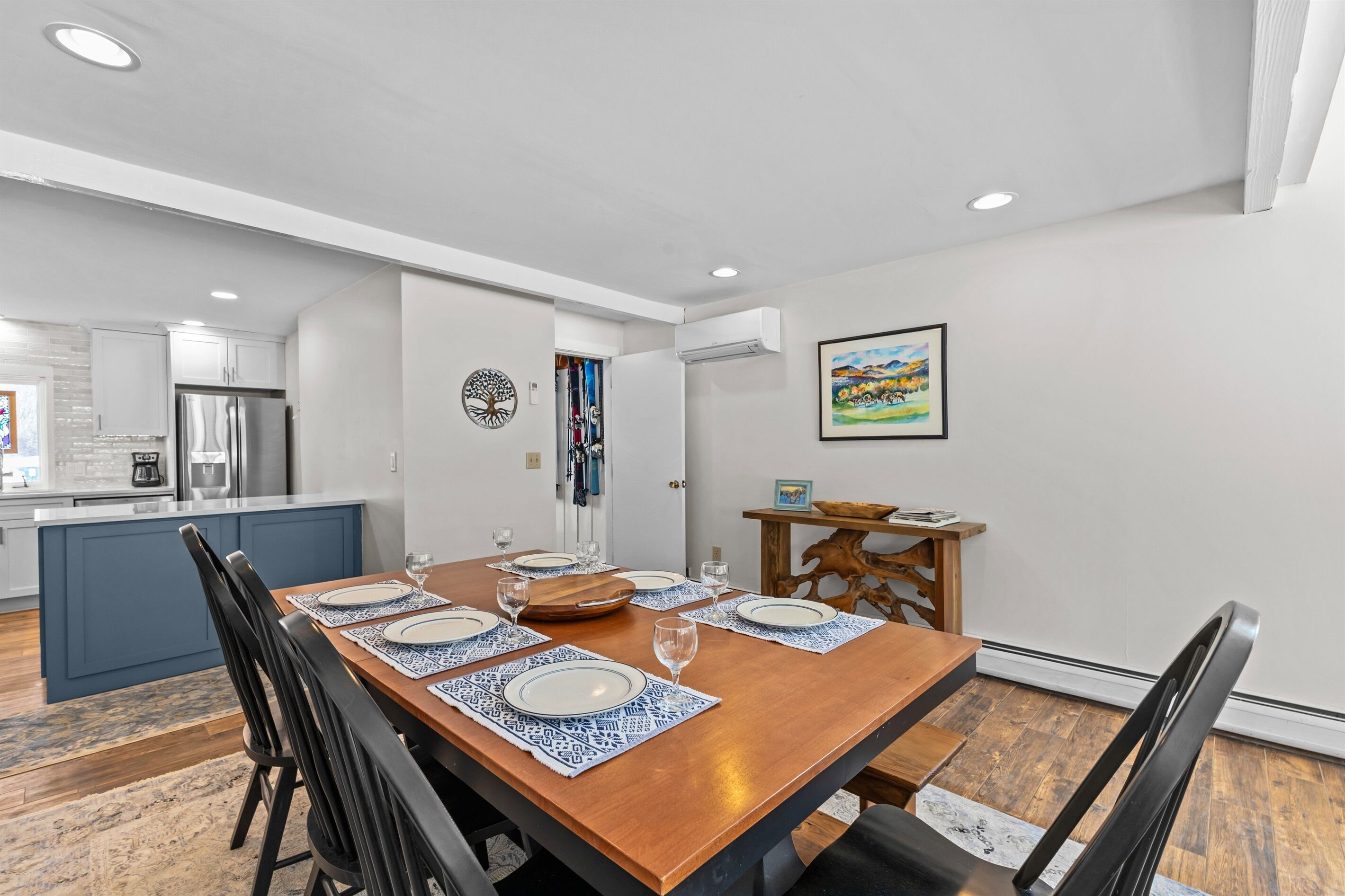 Un comedor con una mesa de madera dispuesta para cuatro, una cocina al fondo y un cuadro en la pared.
