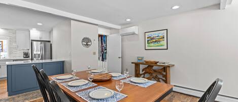 Un comedor con una mesa de madera dispuesta para cuatro, una cocina al fondo y un cuadro en la pared.