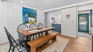 A dining area with a wooden table and chairs, a painting on the wall, and a view into the kitchen.