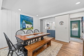 A dining area with a wooden table and chairs, a painting on the wall, and a view into the kitchen. - Charming, sunny 3‑bedroom Village Green Condo (Stowe)