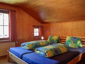 A wooden cabin room with a bed, two pillows, a window with curtains, and a radiator.