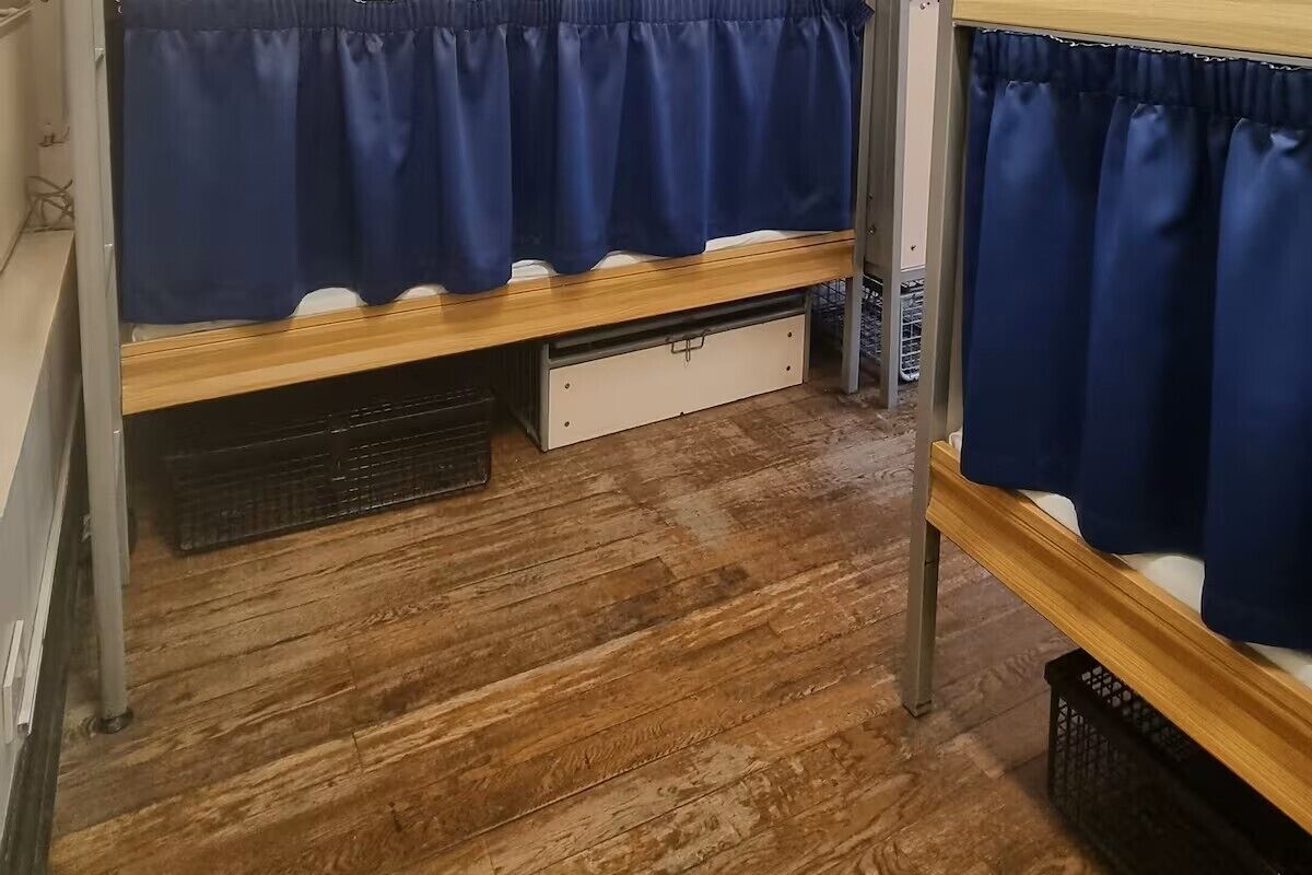 Una cama litera con estructura de madera, un cajón blanco debajo y una cortina azul.