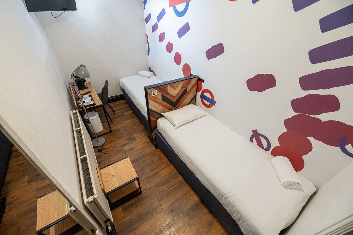 A small, compact room with a bed, a desk, a chair, and a wall with abstract art.