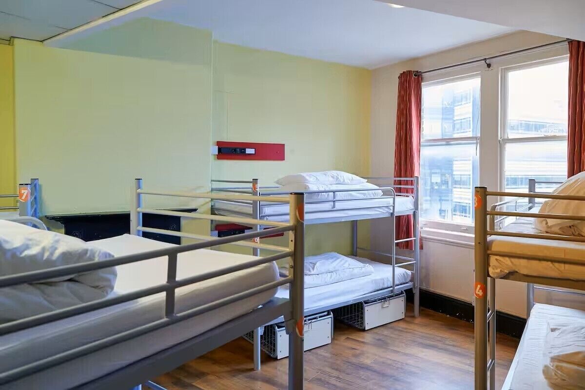 A dormitory room with bunk beds, a red emergency call button, and a window with curtains.