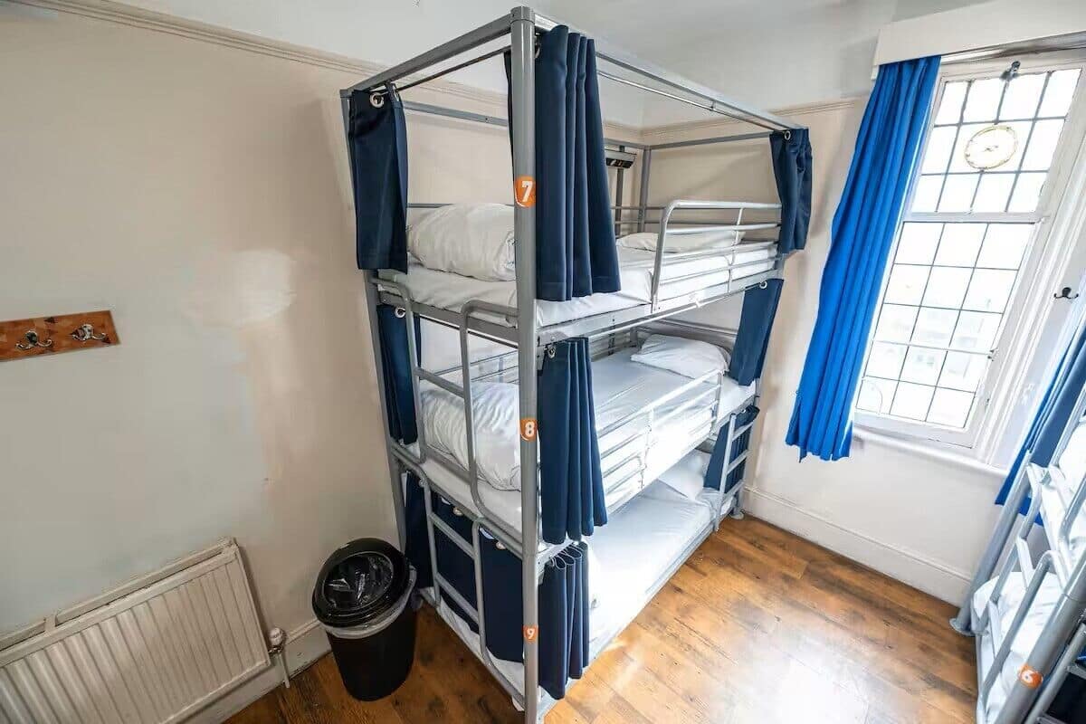 A bunk bed room with a radiator, a window with curtains, and a trash bin.