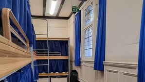 Um dormitório com beliches, cortinas azuis, sinalização de saída de incêndio, uma janela com grades e uma lixeira verde.