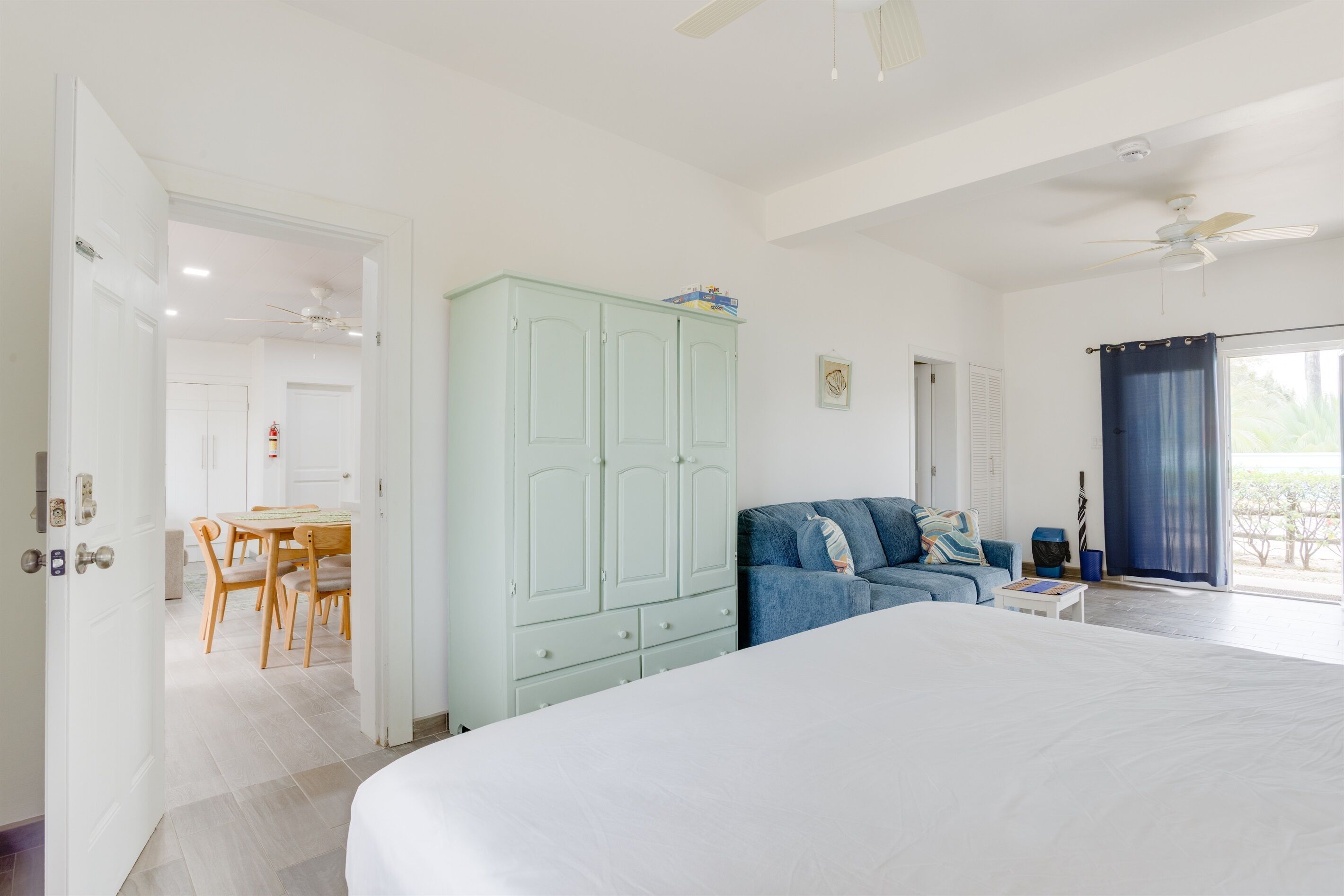 Une chambre avec un grand lit, une armoire bleu clair, un canapé bleu, un coin repas avec des chaises et une table, et un ventilateur de plafond.