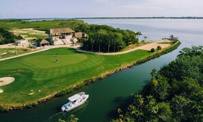 A golf course with a water body, a boat, and a building with a thatched roof. - Spacious 2BR/2BA Placencia Apartment-Pool-Parking (Placencia)