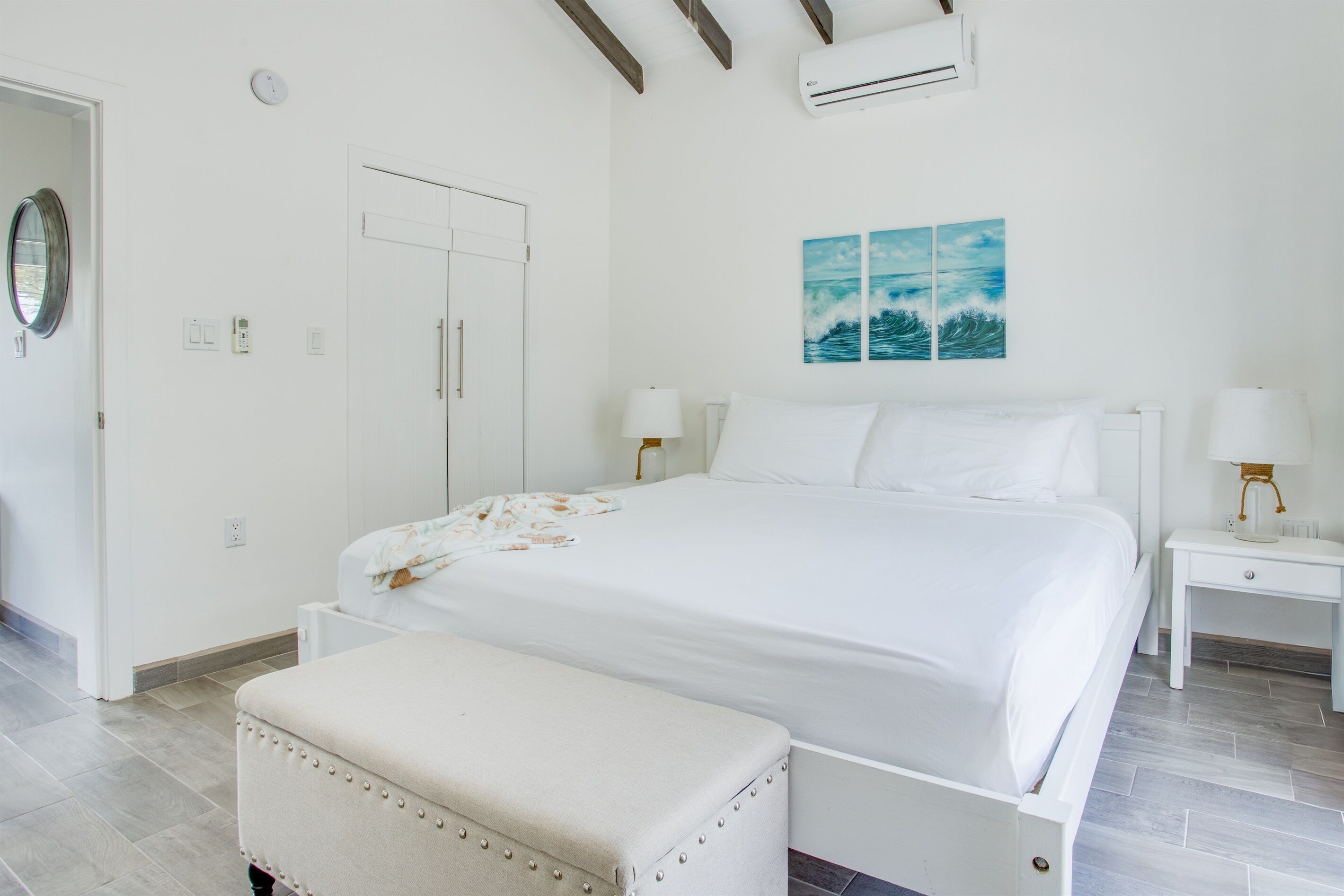 A bedroom with a bed, bedside tables, a bench, a door, and a wall-mounted air conditioner.