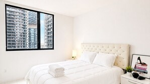 Een moderne hotelkamer met een groot bed, nachtkastje, lamp en uitzicht op hoogbouw.
