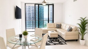 Een moderne woonkamer met een glazen tafel, een beige bank en een groot raam met uitzicht op de stad.