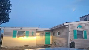 Una casa de dos pisos con una puerta y contraventanas verdes, un pequeño garaje adosado y una antena parabólica visible.