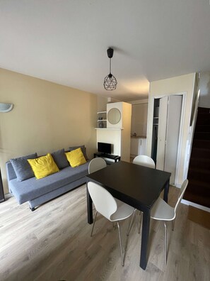 Ein modernes Wohnzimmer mit einem grauen Sofa, weißen Sesseln, einem schwarzen Esstisch und einem kleinen Fernseher.
