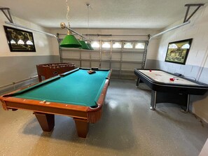 Una mesa de billar con superficie de fieltro verde, una mesa de fútbol de mesa y una mesa de shuffleboard.