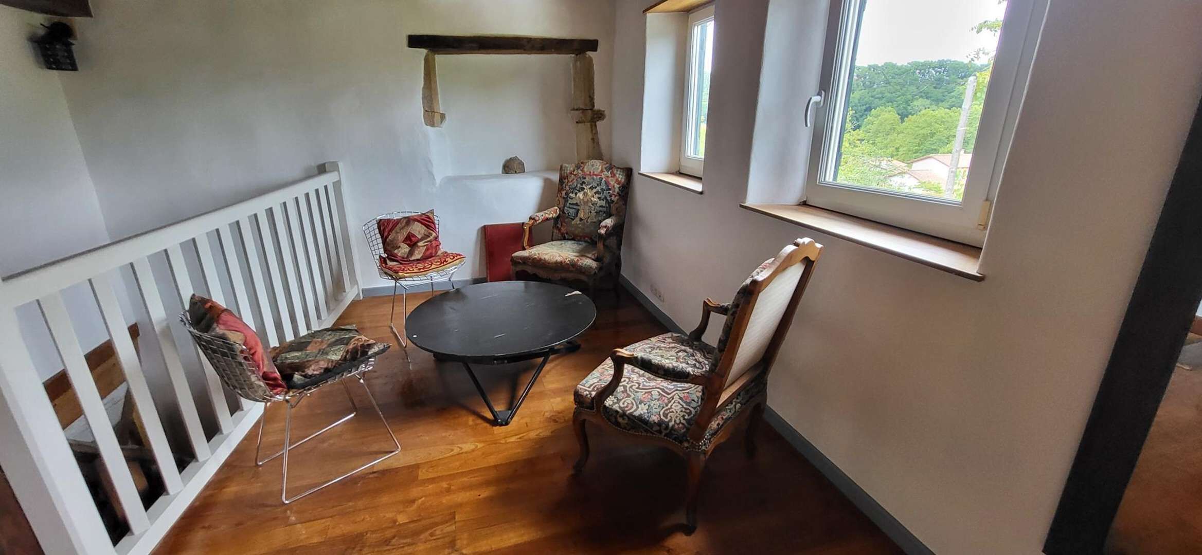 A room with a wooden floor, a round table, two patterned chairs, a white railing, and a window with a view of greenery.