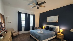 Ein Schlafzimmer mit Bett, Nachttischlampen, Deckenventilator, Nachttisch, Stuhl, Schreibtisch, einem Gemälde und einem Fenster mit Vorhängen.