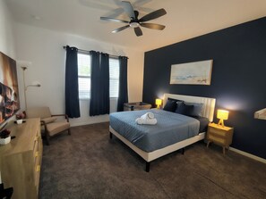 A bedroom with a bed, bedside lamps, a ceiling fan, a nightstand, a chair, a desk, a painting, and a window with curtains. - Family Friendly House Near Disney And Universal (Haines City)
