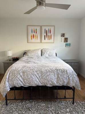 A bedroom with a bed, a ceiling fan, a nightstand with a lamp, and wall art. - 13 Houses to the Strand (Manhattan Beach)