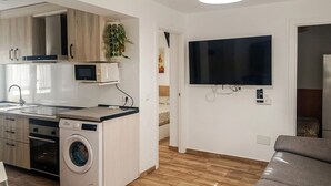 Et lille køkken med mikroovn, ovn og vaskemaskine, et fladskærms-tv monteret på væggen, og en grå sofa i opholdsstuen.
