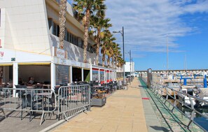 Un café frente al mar con terraza, un puerto deportivo al fondo y un cielo despejado.