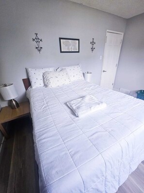 Ein ordentlich bezogenes Bett mit weißer Bettdecke und Kissen, ein Nachttisch mit Lampe und ein gerahmtes Zertifikat an der Wand.