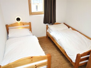 Ein kleines Zimmer mit zwei Holzbetten, weißer Bettwäsche, einem Fenster mit Vorhang und einer Wandleuchte.