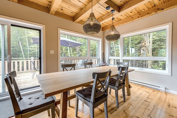 A dining area with a wooden table and chairs, large windows with a view of trees, and a wooden ceiling.
