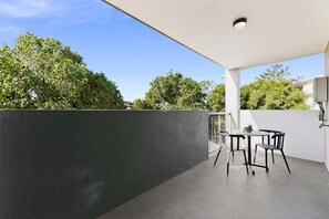 Um pátio ao ar livre com mesa e cadeiras, uma parede escura e árvores ao fundo.