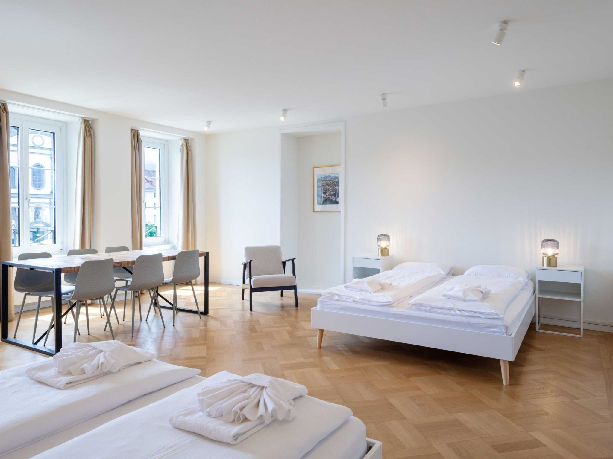 Ein Hotelzimmer mit einem großen Bett, einem Esstisch mit Stühlen, einem Sessel, einem Nachttisch mit Lampe und einem Fenster mit Vorhängen.