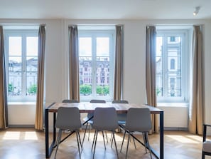 A dining area with a table and chairs, large windows with curtains, and a view of buildings outside. - Apartment  by Interhome (Lucerne)