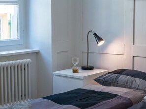 A bedroom with a bed, a bedside table with a lamp, an hourglass, and a radiator. - Apartment  by Interhome (Zurich)