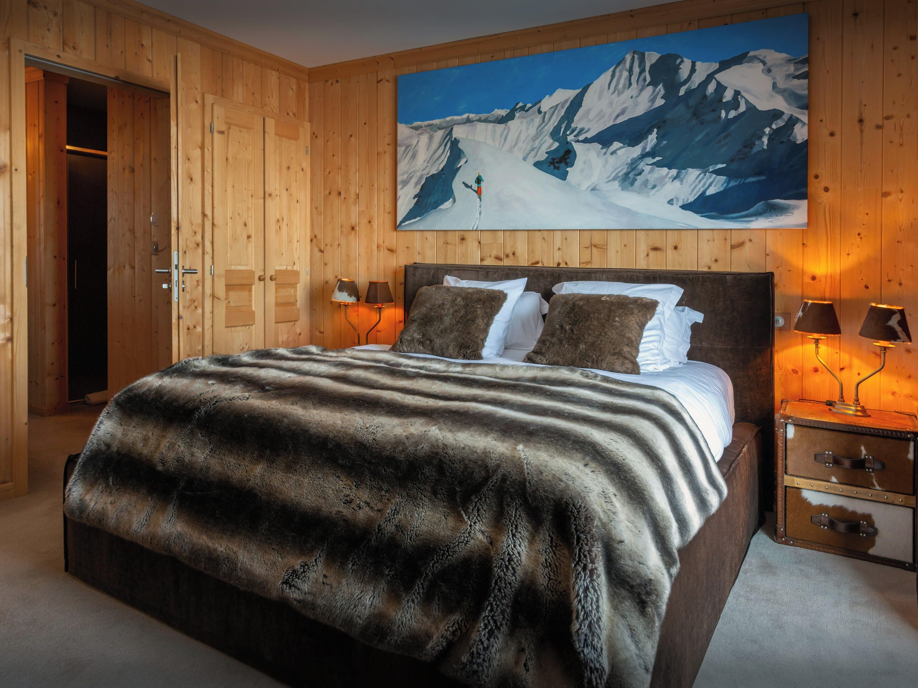 A wooden-paneled room with a bed, two bedside lamps, a wooden nightstand, and a painting of a snowy mountain landscape.