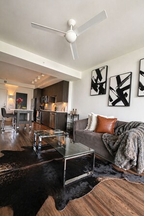 A modern living room with a glass coffee table, a leather sofa, abstract wall art, and a ceiling fan.